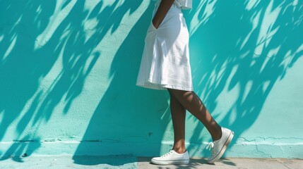 Wall Mural - A woman in a white dress with a high slit standing against a turquoise wall with her hand on her hip casting a shadow.