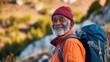 © Z A C - An elderly man with a white beard and a red beanie wearing an orange jacket carrying a blue backpack standing on a mountain with a smile on his face looking at the camera.