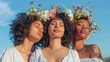 Three women with closed eyes wearing white tops adorned with floral crowns standing against a blue sky exuding a serene and harmonious vibe.