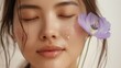A close-up of a woman's face with closed eyes a serene expression and water droplets on her skin with a purple flower gently touching her cheek.