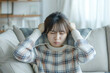 © AI_images - Dissatisfied and angry Japanese young woman sitting on sofa at home and covering ears with pillows from excessive noise