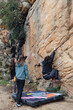 © ADDICTIVE STOCK - Three friends engage in a bouldering session on a rugged mountain face, with one climber ascending as the others spot, safety crash pad in place