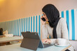© Cultura Creative - Young woman in hijab working on tablet and talking on phone in cafe