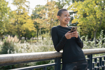 Wall Mural - Smiling athletic woman with smart phone on footbridge