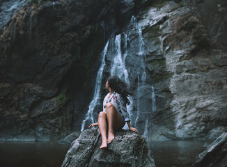 Wall Mural - Portrait of a young woman sitting on a log and looking at a waterfall.