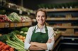 © NOWO - Portrait of  young sales assistant standing in a grocery store, supermarket, in front of shelves with fruits and vegetables. Female store clerk in apron smiling, looking at camera.