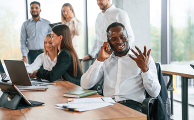 Wall Mural - Black man is talking by the phone. Group of office workers are together indoors