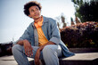 © deagreez - Photo of cheerful cool satisfied youngster with afro hairdo dressed jeans jacket headphones sitting on platform relaxing in park outdoors