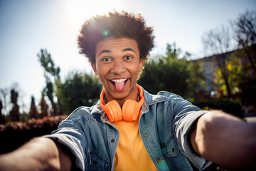 Canvas Print - Photo of funky carefree childish youngster dressed jeans jacket with headphones on neck making selfie stick out tongue in park outdoors