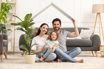 Wall Mural - Housing concept. Happy husband holding plastic roof over his family while sitting on floor at home