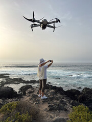 Canvas Print - Rear view of senior woman on a rocky beach taking pictures of the ocean waves, a drone device flights above the lady.  Concept of relaxed elderly person admiring the power of nature