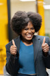 © JoseIMartin - Smiling businesswoman with afro hair gesturing thumbs up in a corporate setting, exuding positivity.