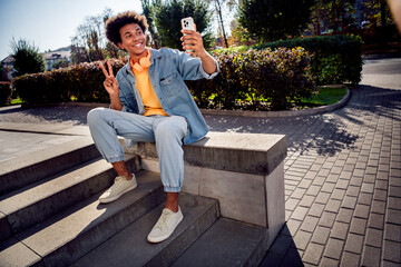 Canvas Print - Photo of optimistic nice youngster dressed jeans jacket headphones sitting on platform show v-sign doing selfie on smartphone outdoors