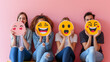 people are sitting side by side holding up large emoji face placards in front of their faces against a pink background.