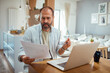 © Davor - Concerned man reading documents and using laptop in kitchen