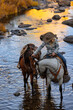 Wyoming Cowgirl in a rocky stream riding a grey horse in the fall working at a ranch leading a horse