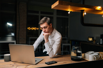 Focused freelancer male using laptop at office workplace. Overworked office worker working on project all night looks at computer screen feeling tired, sleepy after having overwork project overnight