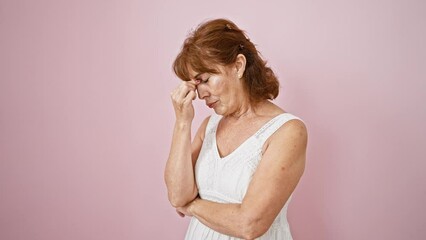 Sticker - Exhausted middle-age woman in dress touches fatigued eyes, rubbing pained nose. stress-induced headache evident. standing in frustration against a solo pink backdrop.