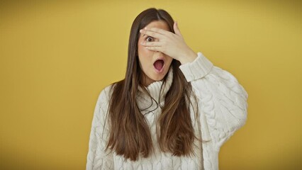 Poster - Shocked young hispanic woman hiding her beautiful face behind fingers, peeking through hand-cover over isolated yellow wall