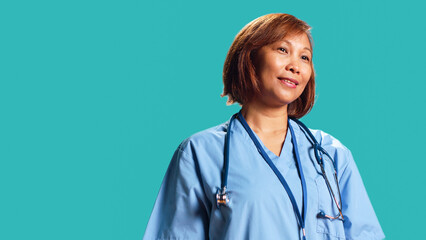 Wall Mural - Happy proficient BIPOC nurse smiling while looking at camera. Portrait of cheerful asian healthcare specialist wearing professional scrubs, isolated over blue studio background