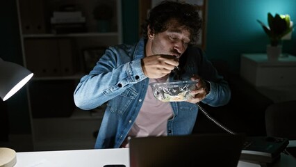 Poster - Hispanic man in jean jacket works late in office, talking on phone and eating salad.