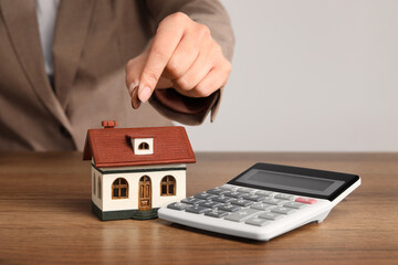 Wall Mural - Mortgage concept. Woman putting coin into house model at wooden table, closeup with space for text