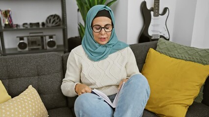 Canvas Print - A mature woman wearing glasses and a hijab reads a book in a cozy living room with a guitar in the background.