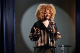 Fototapeta  - Young African American stand up comedian in wig speaking in microphone during performance while standing on stage in front of audience