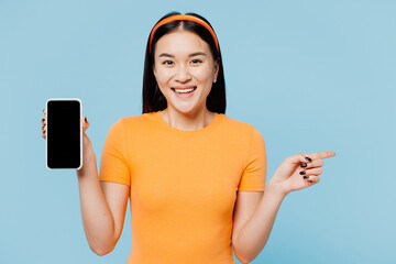 Wall Mural - Young woman of Asian ethnicity wear orange t-shirt casual clothes hold use blank screen workspace area mobile cell phone point finger aside isolated on plain pastel blue background. Lifestyle concept.