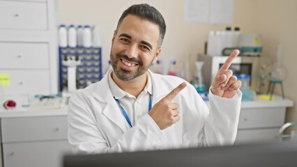 Sticker - Smiling young hispanic man in scientist uniform joyfully points to the side at the lab, confidently presenting with both hands while working on the computer