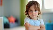 © iuricazac - Young child with arms crossed frowning standing in a room with a green wall and a blurred background that includes a window and a piece of furniture.