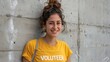 © iuricazac - Young woman with curly hair smiling wearing a yellow t-shirt with the word 'VOLUNTEER' on it standing against a concrete wall.