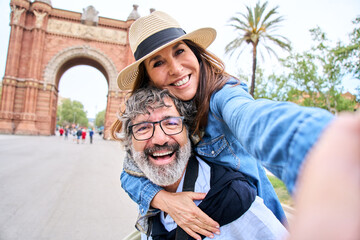 A senior gray hair man wearing a sun hat is giving a piggyback ride to a mature woman in a happy gesture while taking a selfie in touristic city. They seem to be having fun during their leisure travel