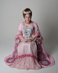 Wall Mural - Fun length portrait of female model wearing an opulent pink gown, costume of a historical French baroque nobility, style of Marie Antoinette. Sitting pose on throne. Studio background.