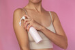 A young woman in beige underwear applies a white moisturizing and nourishing cream to the clean skin of the body using a bottle with a dispenser. Isolated on a pink background