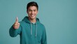 Casual young man in teal hoodie giving thumbs up, with a soft blue backdrop.