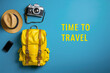 Mochila de color amarillo junto a cámara de fotos, sombrero de paja y teléfono móvil, sobre fondo azul con la inscripción Time to Travel