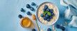 A bowl of oatmeal with blueberries and honey. A spoon and a napkin on a table. A jar of honey and a blueberry on a light blue background Top view space to copy.