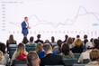 A professional presenter addresses an attentive audience at a business conference with focus on data charts.