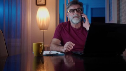 Wall Mural - Enjoying good working day. Confident mature man working on laptop and talking on the mobile phone while sitting at his working place in home office. 4k video footage