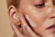 Close up highly detailed shot of a young woman applying cream on her freckled skin. Cropped shot of woman applying moisturizer on cheek with fingers.