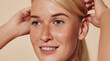 © Artem Varnitsin - Close up highly detailed portrait of a young smiling woman with freckles. Cropped image of a beautiful blond female adjusting hair against a beige backdrop.