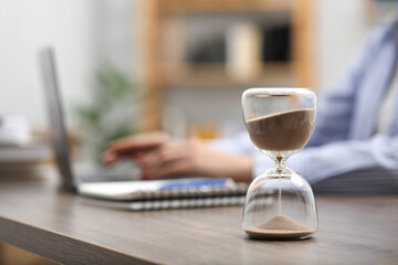 Wall Mural - Hourglass with flowing sand on desk. Woman using laptop indoors, selective focus