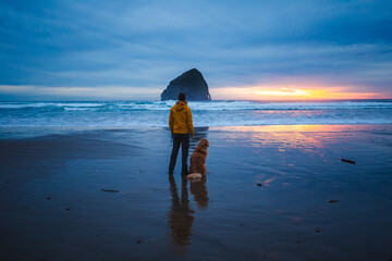 An image of a man with his dog standing on Pacific City Beach at sunset on the Oregon coast, with Chief Kiwanda Beach on the background