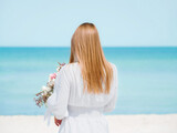 Fototapeta Desenie - Joyful Young Woman Holding a Bouquet of Flowers on a Beach with Blue Sky Background