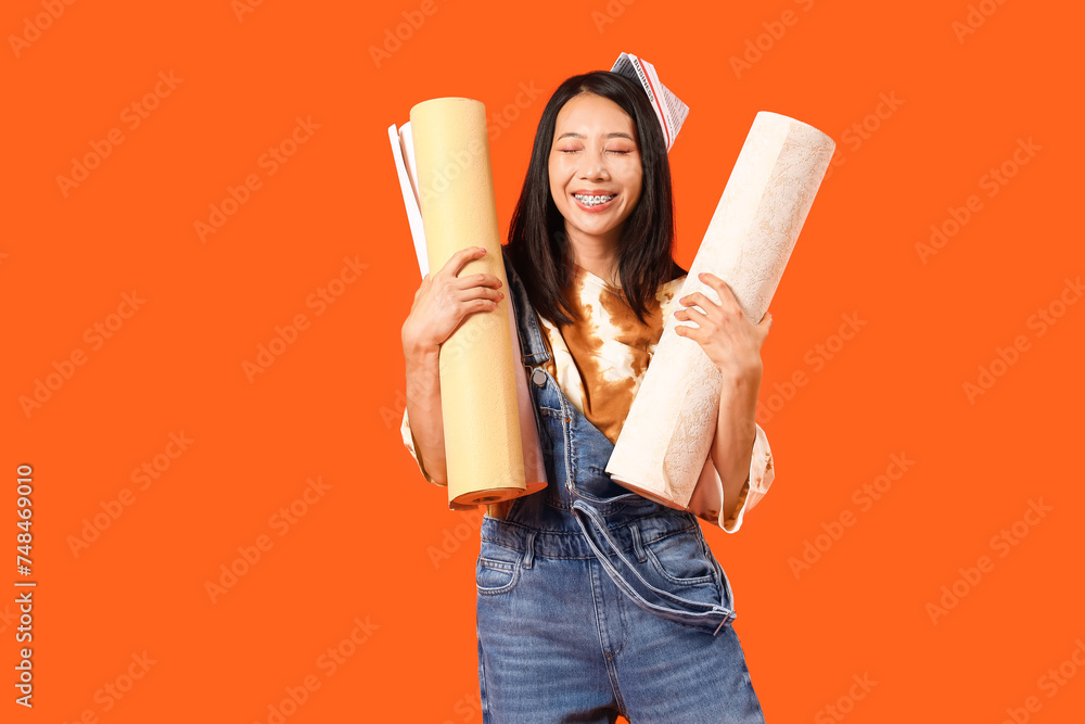 Young Asian woman with wallpaper rolls on orange background