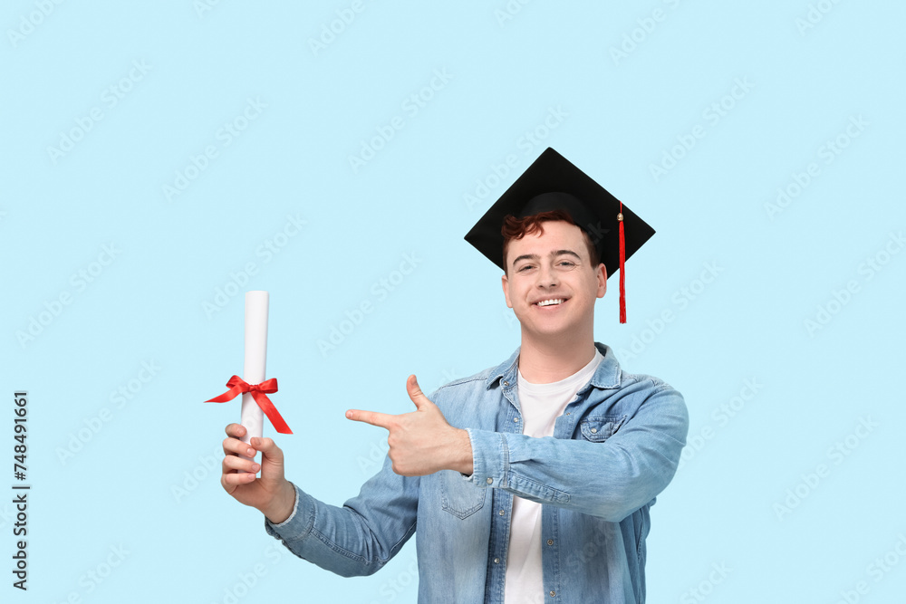 Male student in mortar board pointing at diploma on blue background