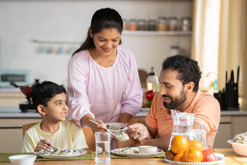 Happy Indian woman serving food to her son and husband on dining table at home - concept of family bonding, household responsibility and togetherness