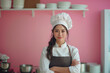 Portrait of young Asian woman in chef hat and apron standing with arms crossed in kitchen.