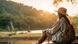 A young woman wearing a Hat, a Backpack is sitting on the riverbank in the morning at dawn, admiring the Beauty of nature. Horizontal Banner, Copy Space, Tourists, Travel, Vacations, Summer concepts.